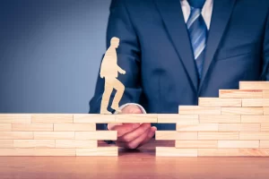 A businessman in a navy blue suit and striped tie holds a wooden block to bridge a gap between two stacked sets of wooden blocks. A wooden cutout figure of a person is shown walking across the bridge towards the ascending steps. The image symbolizes support, guidance, personal development, career growth, and overcoming obstacles.